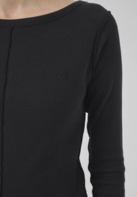 Close-up of a person wearing a black ribbed long-sleeve shirt with a small embroidered logo on the left chest.