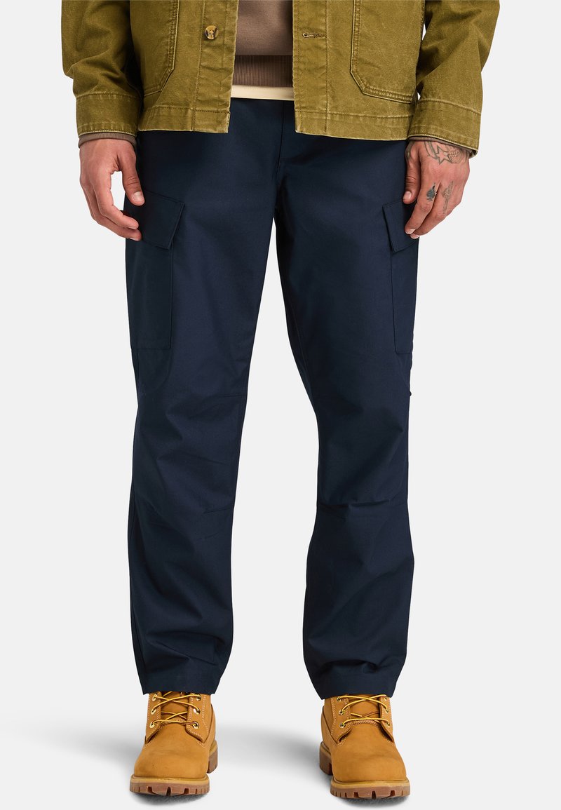 Marine cargobroek met zijzakken, gemaakt van duurzaam materiaal, gecombineerd met tan laarzen. De outfit bevat een groene jas over een beige top.