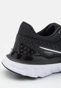 Chaussure de course noire en Flyknit avec un dessus tissé texturé, un contrefort de talon noir brillant et une semelle intermédiaire blanche avec un motif ondulé.