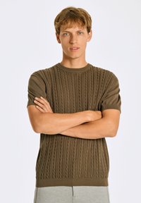 Brun stickad tröja med ett texturerat flättpatent, korta ärmar och rund halsringning, bärs av en modell med armarna korsade.