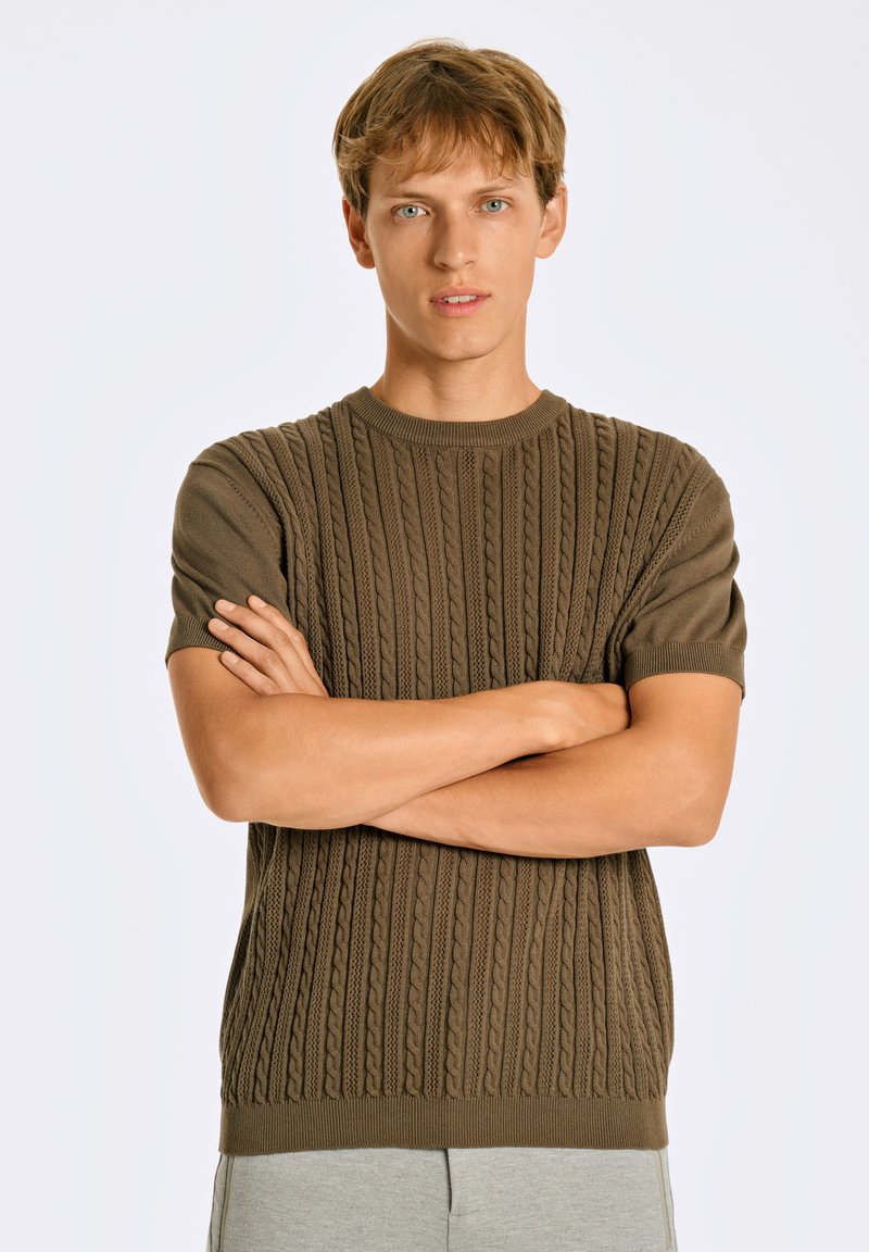 Brun stickad tröja med ett texturerat flättpatent, korta ärmar och rund halsringning, bärs av en modell med armarna korsade.