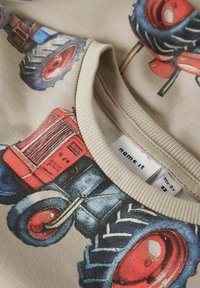T-shirt di cotone beige chiaro con illustrazioni colorate di trattori, colletto a coste e etichetta in tessuto che mostra i dettagli del marchio e della taglia.