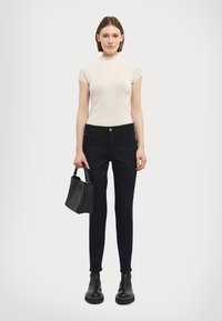 Light beige ribbed top with high neckline, paired with fitted black jeans and black ankle boots. Model holds a small black handbag.