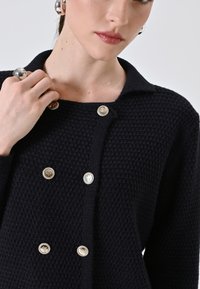 Cardigan a maglia navy con un motivo testurizzato, colletto rotondo e quattro bottoni metallici. Caratterizzato da un design pulito e apertura frontale.
