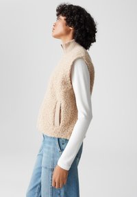 Maglione sans maniche beige con texture, colletto alto a coste, tasca laterale, indossato sopra una maglietta bianca a maniche lunghe, abbinato a jeans blu.