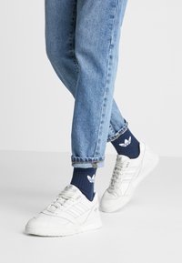 Des baskets de sport blanches avec des détails texturés et trois rayures sur le côté, portées avec des chaussettes bleues marine ornées d'un logo blanc, assorties à un jean en denim clair retroussé.