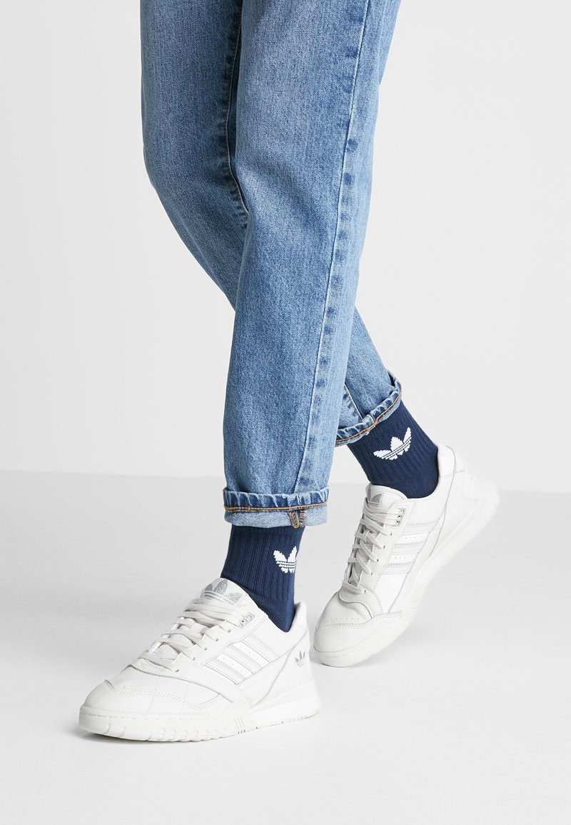 Des baskets de sport blanches avec des détails texturés et trois rayures sur le côté, portées avec des chaussettes bleues marine ornées d'un logo blanc, assorties à un jean en denim clair retroussé.