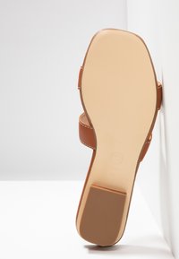Brown leather sandal with a smooth sole, minimal strap design, and visible white stitching. The outsole is flat with textured grip.