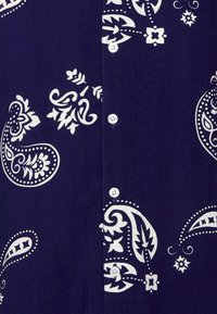 Námořnická košile s velkými bílými paisley a květinovými vzory. Přední zapínání na čtyři viditelné knoflíky, hladká textura látky.