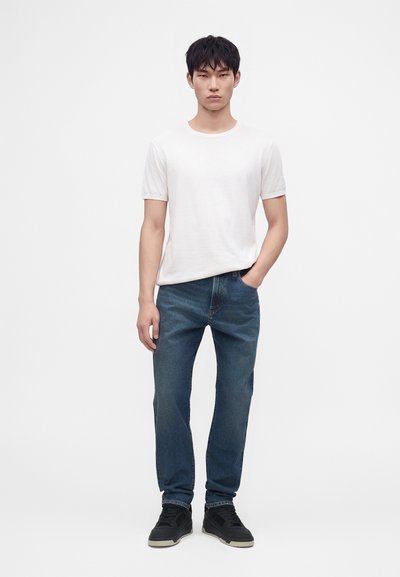 Jeune homme debout en tenue décontractée avec chemise blanche à manches courtes, jean bleu, baskets noires, une main dans la poche, sur fond uni.