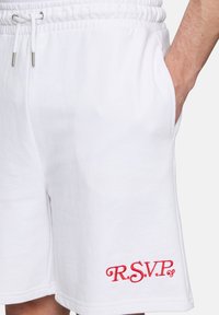 Shorts en coton blanc avec une taille élastique, présentant un logo "R.S.V.P" brodé en rouge sur le côté gauche et des poches latérales.