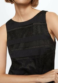 Haut noir sans manches présentant des panneaux en dentelle texturée et des bandes transparentes, avec un col lisse. Le tissu affiche des motifs complexes.