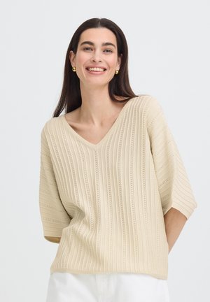 Usmievavá žena s dlhými tmavými vlasmi, ktorá nosí zlaté kruhové náušnice a krémový žebrovaný sveter s výstrihom do V a polovičnými rukávmi, pózuje na bielom pozadí.