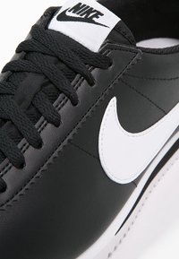 Baskets noires Nike avec logo swoosh blanc, lacets noirs et étiquette blanche affichant le texte Nike noir sur la languette.