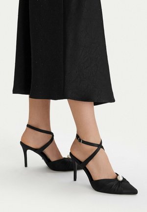 Tacones altos negros de punta afilada con tiras en el tobillo y una perla decorativa en el frente, usados con una falda midi negra.