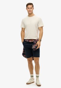Beige bomulds T-shirt, navy shorts, rutet skjorte bundet om livet, hvide sneakers med brune såler og sokker med grønne accenter.