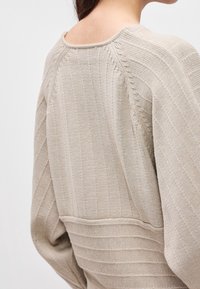 Gros plan d'une personne portant un pull en maille côtelée beige avec des motifs texturés verticaux et horizontaux sur le dos et les manches.