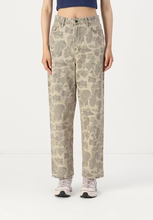 Beige en grijze camouflagebroek met bloemenprint op halfhoge taille, gedragen met lichtroze sneakers en een cropped marineblauw topje, gedragen door een staande persoon.