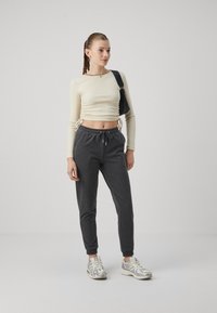 Šviesiai smėlio spalvos ilgas rankovės crop topas su šoniniu raukinimu, derinamas su tamsiai pilkiais džemperiais. Aprangą papildo sidabrinės sportinės bateliai.
