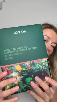 Person hält eine grüne Aveda Moisture Marvels Hautpflegeset-Box mit einem bunten abstrakten Design auf der unteren Hälfte.