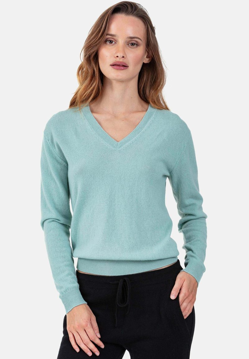 Pull turquoise clair à col en V, fabriqué en tissu doux, avec des manches longues et un ourlet côtelé, porté avec un pantalon noir.