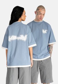 Lyseblå oversized t-shirts i bomuldsstof, hvide detaljer på ærmerne, med et iøjnefaldende grafisk design på ryggen og forsiden.