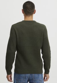 Dunkelgrüner Strickpullover mit strukturierten Mustern auf dem oberen Rücken und glatten Rippbündchen an den Ärmeln und am Taillenbund, kombiniert mit blauen Jeans.