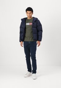 Námorná puffa bunda s kapucňou, prešívaný dizajn a elastické manžety, v kombinácii so zeleným tričkom s logom a tmavými džínsami. Biele tenisky.