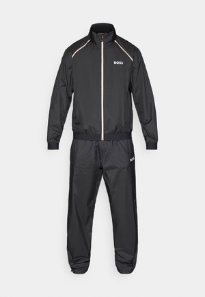 Tuta nera realizzata in materiale leggero, composta da una giacca con zip e colletto alto e pantaloni con elastico in vita abbinati. Accentuata da dettagli dorati.