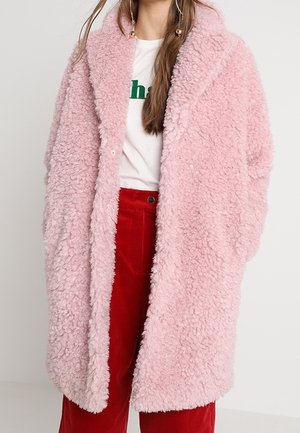 Cappotto di pelliccia sintetica rosa fluffy con colletto a scialle, caratterizzato da una superficie testurizzata e una vestibilità oversize. Indossato sopra una camicia bianca e pantaloni rossi.