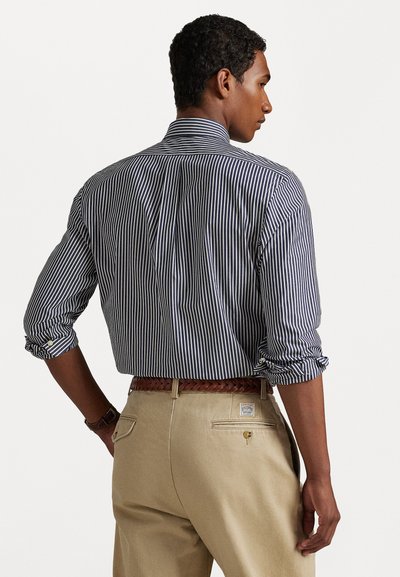 Πουκάμισο με κουμπιά σε ναυτικό μπλε και λευκές κάθετες ρίγες, με ρολό μανίκια, βαμβακερό ύφασμα και κλασικό γιακά, συνδυασμένο με καφέ παντελόνι και πλεχτή ζώνη.