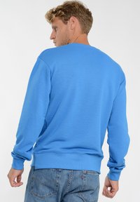 Niebieski sweter z prążkowaną teksturą, okrągłym dekoltem i długimi rękawami. Posiada dopasowany pas i mankiety, w zestawie z jasnoniebieskimi dżinsami.
