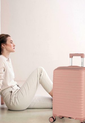 Mujer con ropa beige sentada en el suelo junto a una maleta rosa rígida con asa extendida y ruedas, sobre un fondo liso.