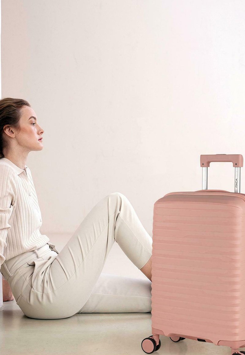 Mujer con ropa beige sentada en el suelo junto a una maleta rosa rígida con asa extendida y ruedas, sobre un fondo liso.