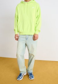 Neon grön hoodie med känguruficka och dragsko, ihopparad med ljus tvättade säckiga jeans och flerfärgade sportskor.