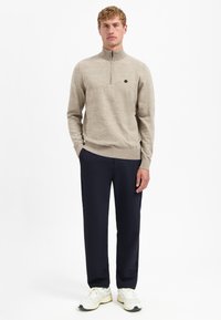 Beige Pullover mit Reißverschluss und strukturiertem Finish, kombiniert mit marineblauen Hosen und weißen Sneakers. Das Outfit zeichnet sich durch ein einfaches, modernes Design aus.