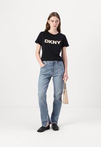 T-shirt noir à manches courtes avec le logo "DKNY" en beige, associé à un jean droit bleu clair et des mocassins noirs ; tenant un sac beige.