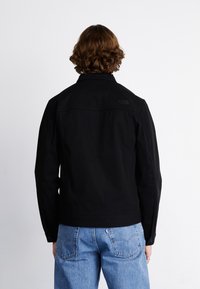 Veste en coton noire avec un col montant et des poignets à boutons. Présente un logo discret dans le dos, associée à un jean en denim bleu clair.