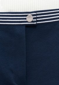 Pantalon bleu marine texturé avec une ceinture rayée bleu marine et blanche ainsi qu'un bouton argenté gravé "Lacoste".
