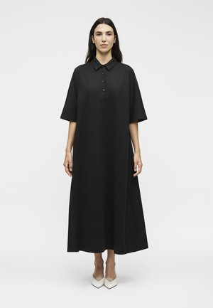 Γυναίκα που στέκεται με το πρόσωπο μπροστά, φορώντας ένα χαλαρό, μαύρο, κοντομάνικο, κουμπωτό μίντι φόρεμα και λευκά μυτερά τακούνια σε απλό φόντο.