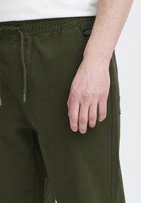 Olívazöld nadrág puha anyagból, gumis derékpánttal és húzózsinórral. Oldalzsebekkel és minimális címkedíszítéssel rendelkezik.