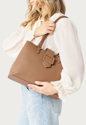 Femme portant un chemisier blanc et un jean clair, tenant un grand sac fourre-tout en cuir marron avec un détail de boucle sur la sangle d'épaule.