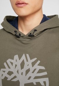 Primo piano di una persona che indossa un hoodie verde oliva con un design astratto di un albero bianco e dei lacci neri regolabili sopra una maglietta blu navy.