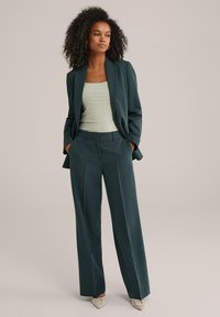 Completo su misura verde scuro con pantaloni a gamba ampia e un blazer strutturato. Indossato con una canotta a coste verde chiaro e tacchi a punta color crema.