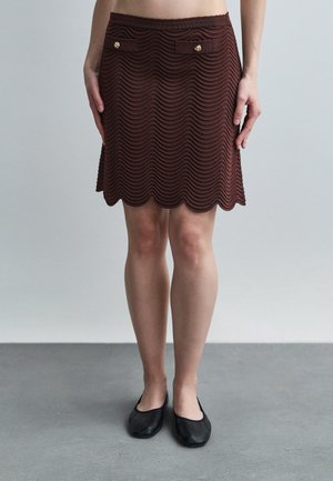 Kobieta ubrana w brązową, teksturowaną spódnicę z falowanym brzegiem i dwoma przednimi kieszeniami na guziki, w połączeniu z czarnymi płaskimi butami na szarym tle podłogi.