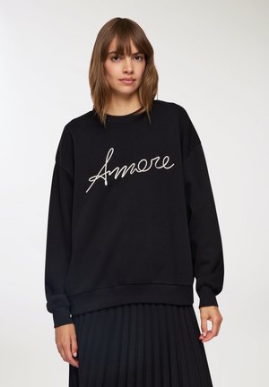 Donna con i capelli castani che indossa una felpa nera con la scritta "Amore" e una gonna plissettata nera, posata su uno sfondo bianco puro.