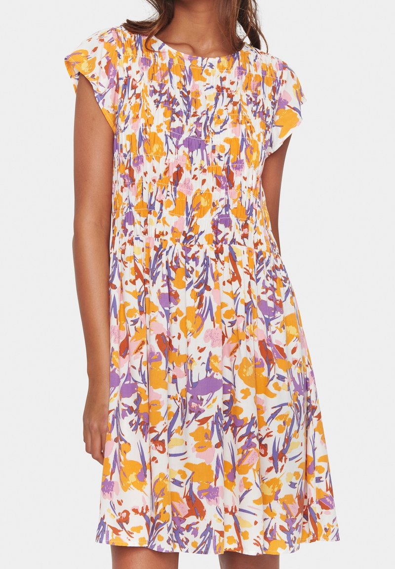 Femme portant une robe sans manches, longueur genou, avec un imprimé floral orange, violet et rose et un détail froncé sur la poitrine.