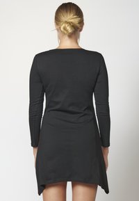 Robe noire à manches longues avec un col rond et un ourlet évasé, présentant une texture lisse et des fentes latérales discrètes pour faciliter les mouvements.