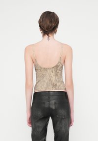 Pessoa com cabelo curto, vestindo uma t-shirt sem mangas de camuflagem bege e calças pretas texturizadas, exibindo uma pequena tatuagem floral na parte superior do pescoço.