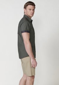 Camisa verde oliva de manga corta con puños enrollados, cierre de botones y bolsillo en el pecho, combinada con pantalones cortos beiges hasta la rodilla. Diseño minimalista.